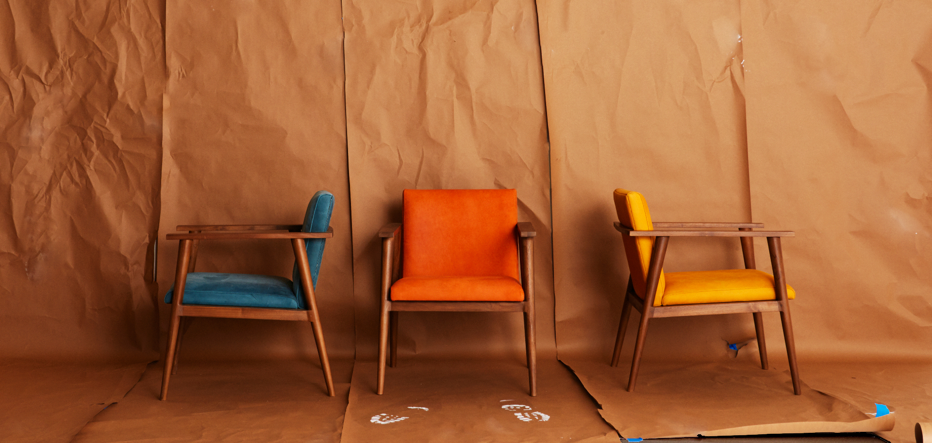 Trio of mid-century walnut armchairs upholstered in teal, orange, and amber against a kraft-paper backdrop