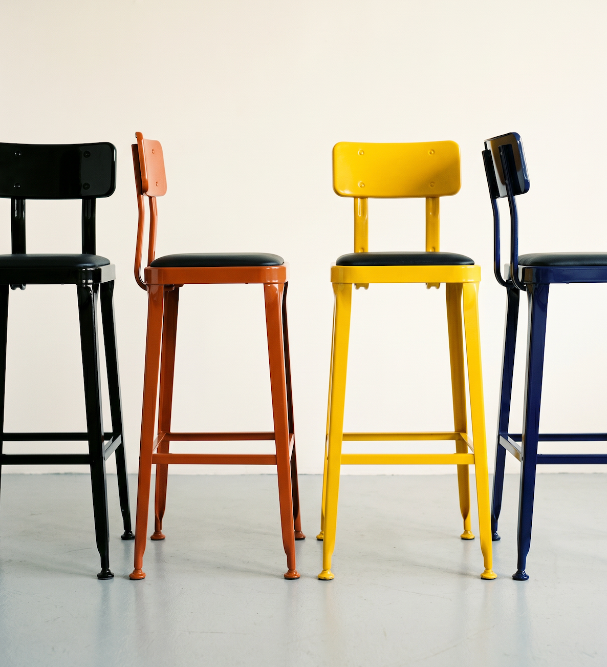A line-up of bar stools in black, oxide orange, sulphur yellow and ink blue against a soft white wall